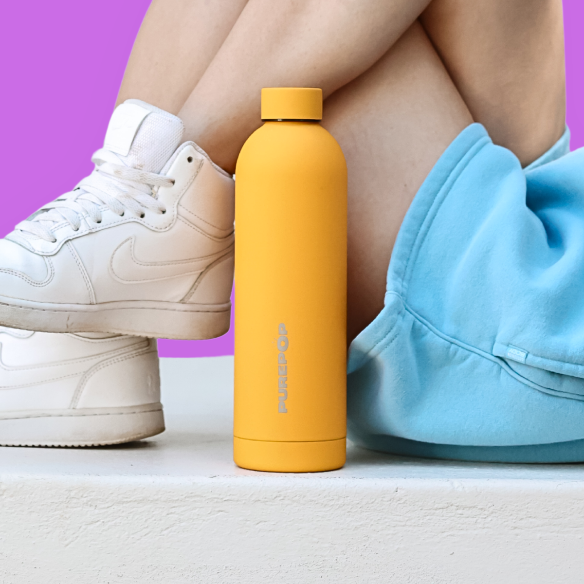 A woman with long blonde hair, wearing a pink cap and red shirt, sits indoors holding a lavender water bottle with Klarissa written on it in gold letters. Her nails are painted light blue.