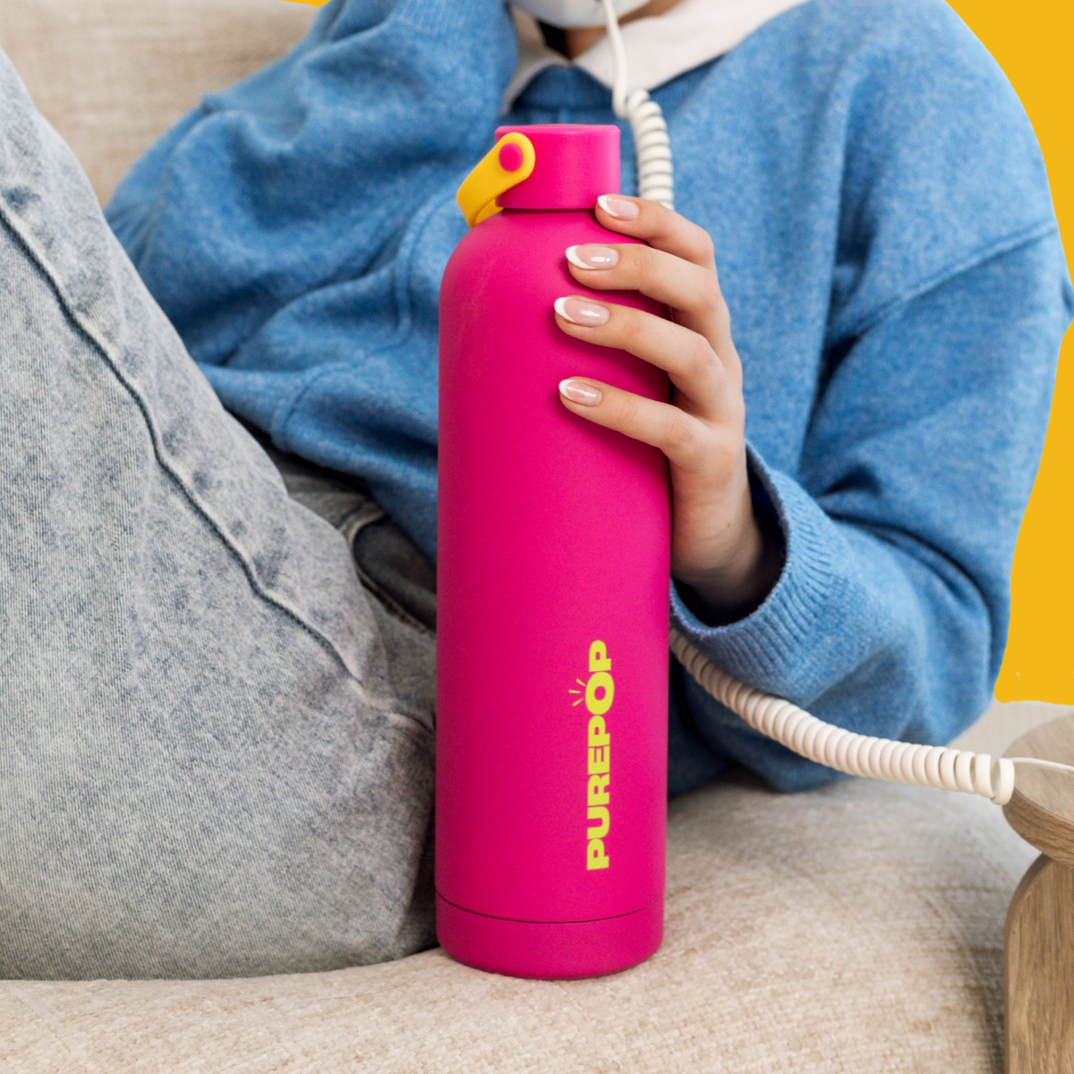 A smiling woman in a red outfit sits on a chair, holding three colorful water bottles—yellow, blue, and green—against a plain white background.