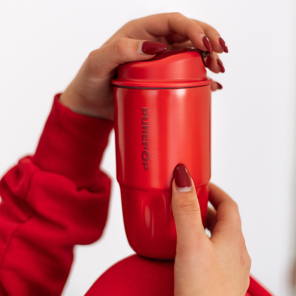 A person with red painted nails and a red sleeve holds a Sip Series | Chili Red reusable coffee cup with the brand PUREPOP visible on the side.