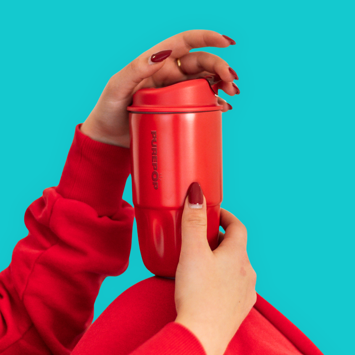 A person with red painted nails and a red sleeve holds a Sip Series | Chili Red reusable coffee cup with the brand PUREPOP visible on the side.