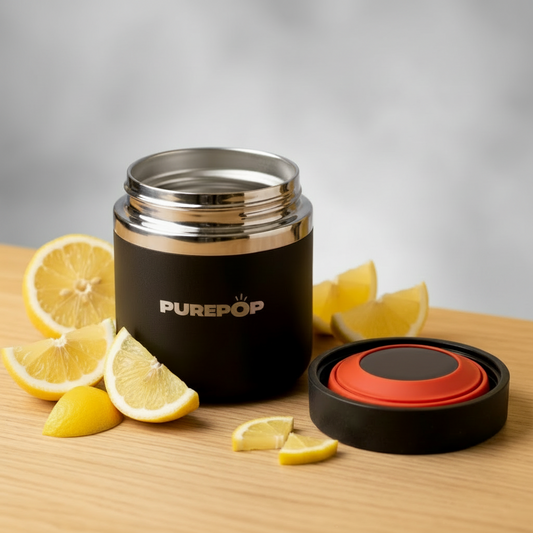 A Purepop Food Flask in Onyx Black with a red and black lid sits open on a wooden surface surrounded by lemon wedges, its modern, minimalist design standing out against a blurred gray background.