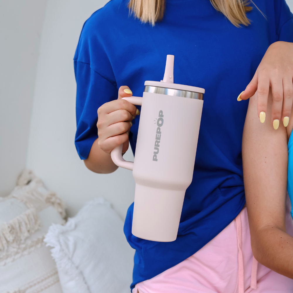 A person in a blue shirt holds a large Carry Series | Rose Quartz insulated travel mug with a straw, standing next to someone in light blue, with pillows in the background.