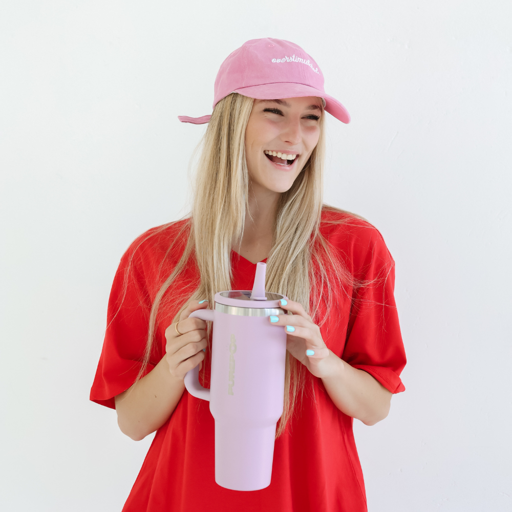 A smiling woman with long blonde hair, in a pink cap and red shirt, holds the Carry Series | Soft Lilac double-walled stainless steel insulated travel mug with a leakproof flip lid and straw against a plain white background.