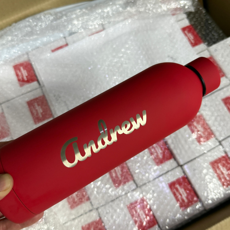 A hand holds a red water bottle with the name Andrew printed on it, above a box filled with bubble-wrapped items and white packages.