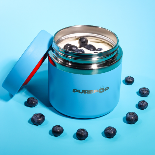 A blue metal PUREPOP container filled with yogurt and blueberries sits open, its lid nearby. Fresh blueberries form a circle around the container on a blue background.