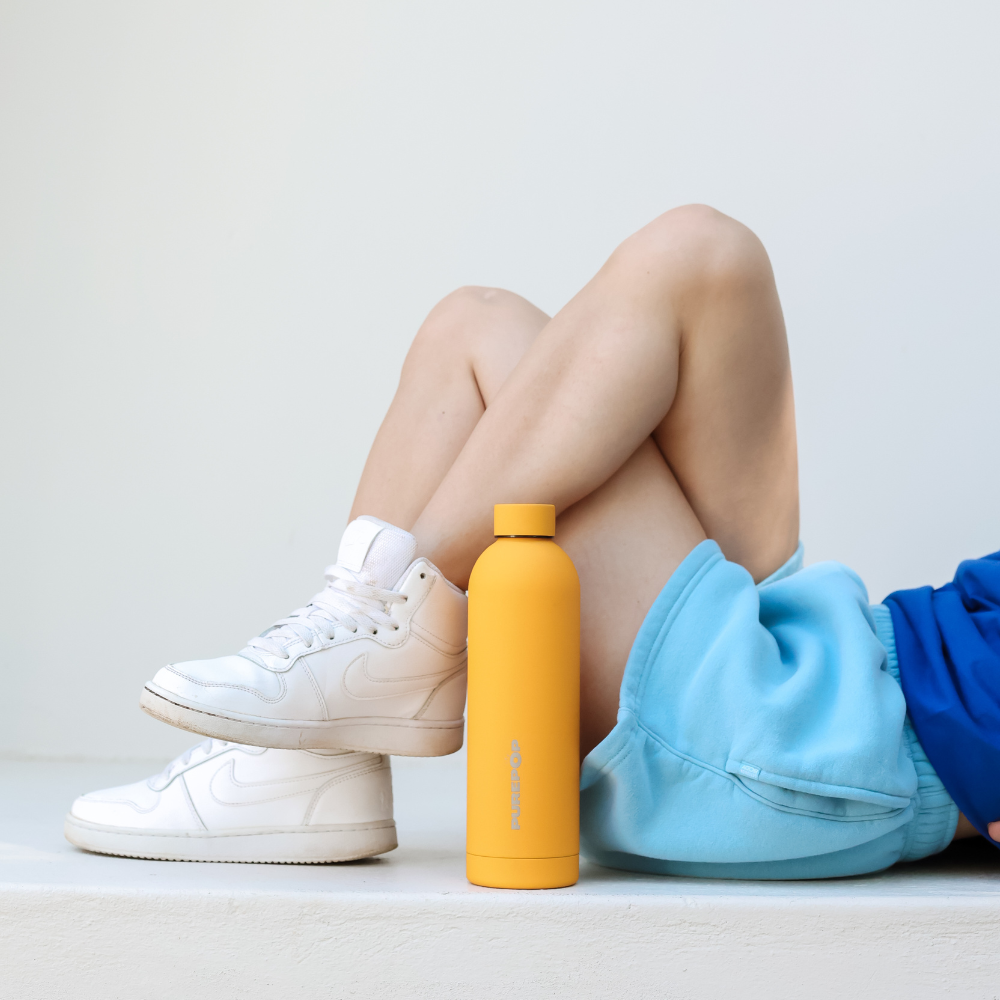 A person in white sneakers and blue shorts lies with bent legs next to the Core Series | Sunshine Yellow leakproof insulated flask, set against a plain white background.