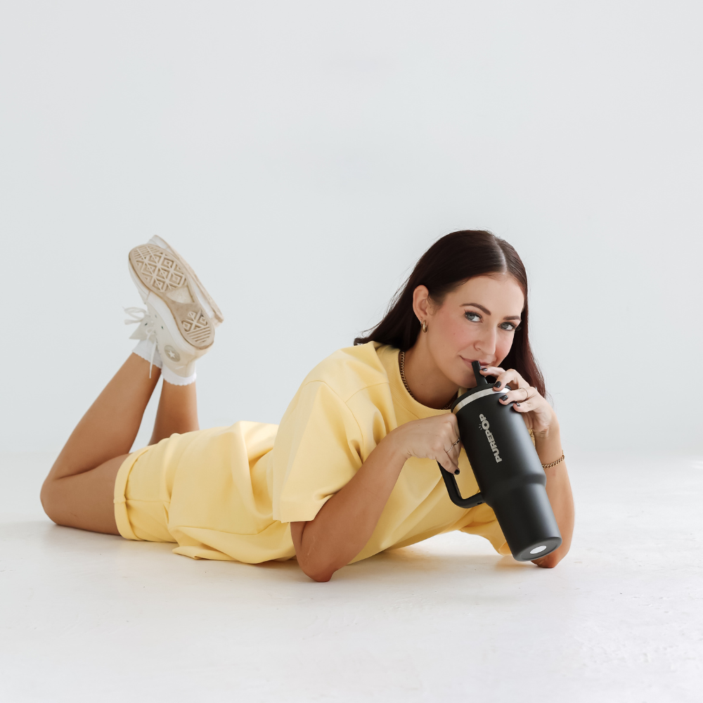 A woman in a yellow outfit, white sneakers, and long dark hair lies on a white floor, smiling and sipping from the Carry Series | Onyx Black double-walled stainless steel tumbler with a leakproof flip lid.