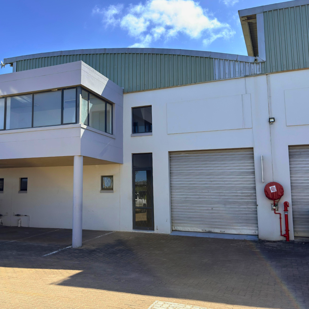 A modern industrial building with a large metal roller door, white walls, glass windows, and a red fire hose reel mounted on the wall. The sky is partly cloudy.