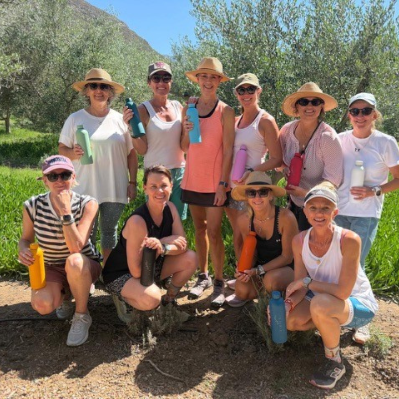 Ten women pose outdoors in a sunny, green area, all wearing hats and sunglasses, each holding a colorful water bottle. They are smiling and dressed in casual, summery clothes.