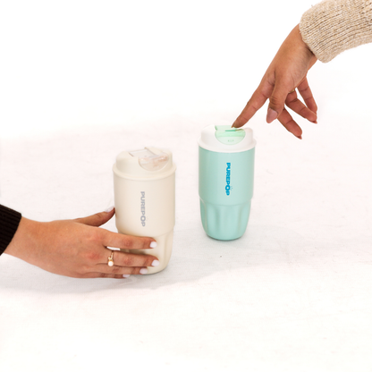 Two hands reach for reusable travel cups—one cream and one Soft Aqua from the Sip Series, labeled PURETOP—placed on a white surface. These stylish Sip Series cups double as a leakproof travel mug and reusable coffee cup, ensuring your drinks stay secure and fresh throughout the day.