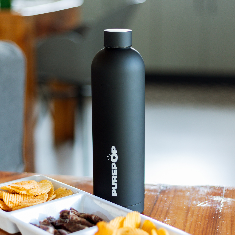 A Core Series Plus | Onyx Black water bottle stands on a wooden table beside trays of potato chips and meat snacks in a bright indoor setting. This matte black insulated flask features double-walled stainless steel construction to keep your drinks at the perfect temperature.