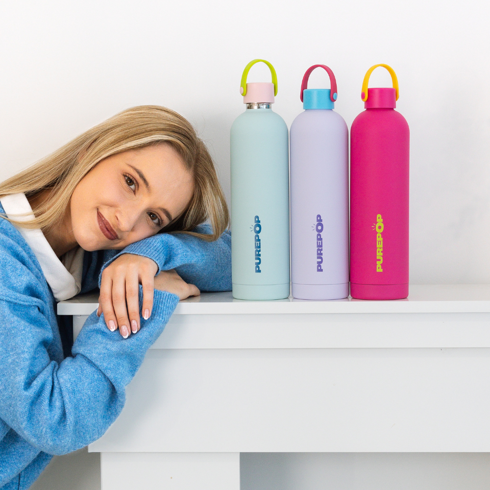A woman in a blue sweater leans her head on her hands beside three colorful Core Series flasks—light blue, lavender, and pink—lined up on a white table against a plain white wall. Each Core Series | Carry Lid | Crimson & Blue flask features a convenient carry lid and a secure, leakproof seal for effortless portability and peace of mind.