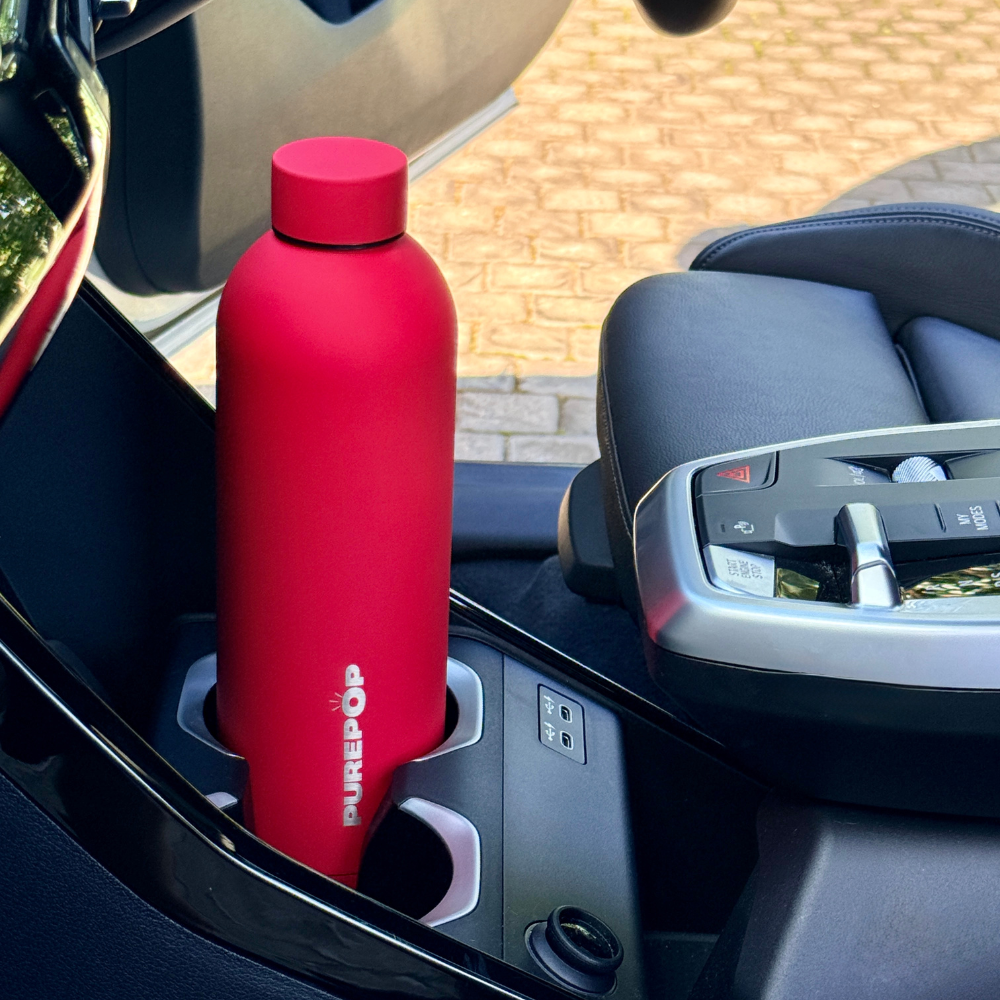 A Crimson Red Purepop Core Series leakproof water bottle sits in the car cup holder beside the gear shift, with part of the driver’s seat and door visible.