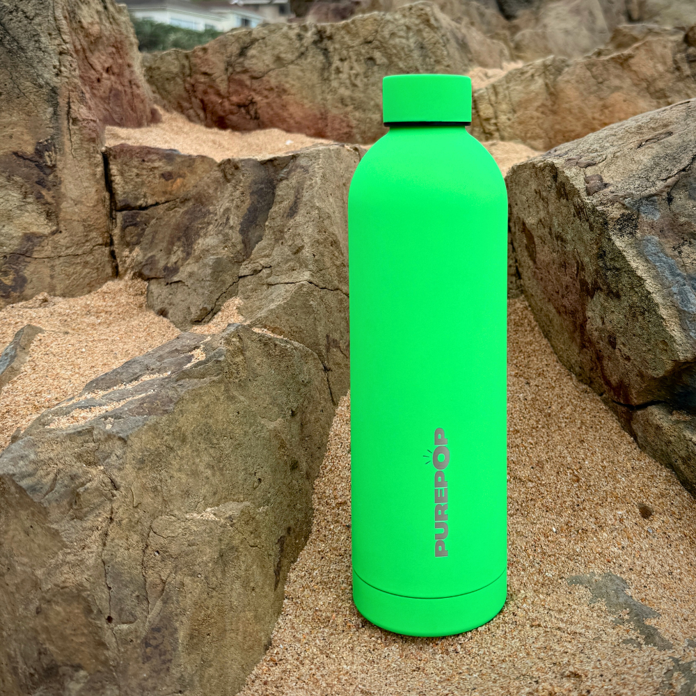 The Purepop Core Series | Electric Green leakproof water bottle stands upright on sandy ground, surrounded by large, rugged rocks.