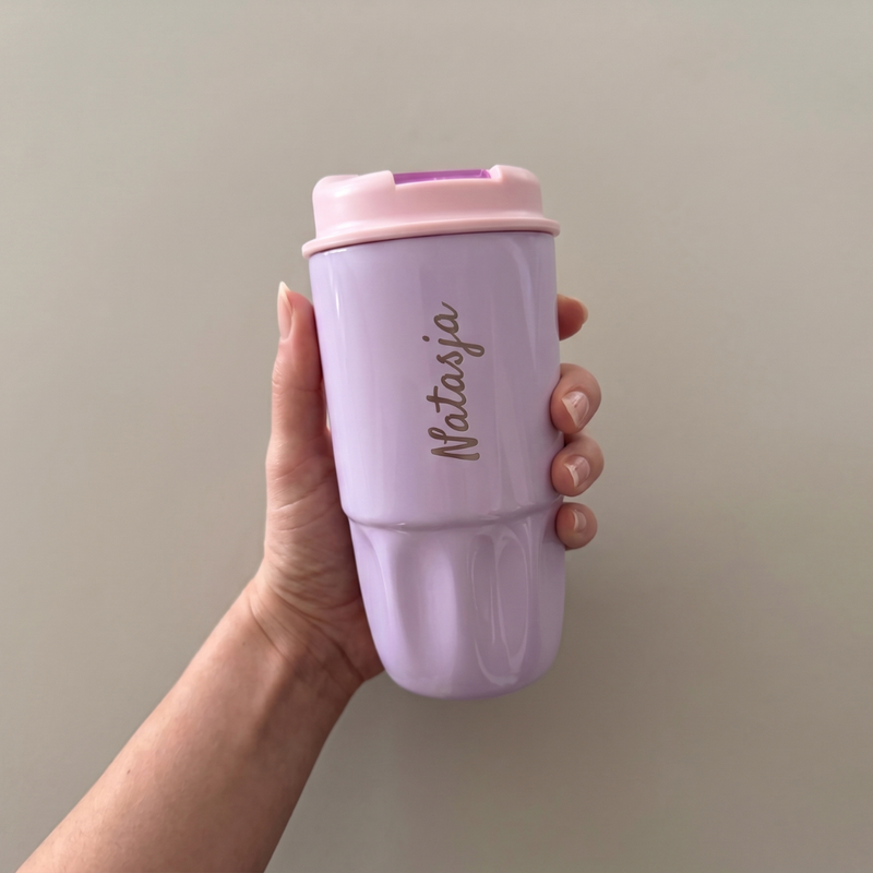 A hand holding a pastel purple reusable cup with a light pink lid. The name Notasja is written vertically on the cup in cursive script. The background is plain and light-colored.