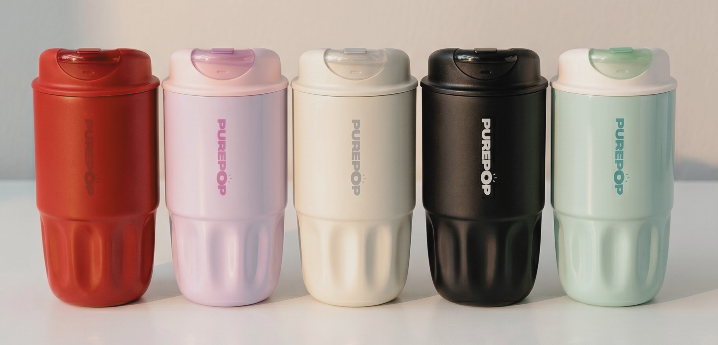 Five PUREPOP reusable tumblers in red, light pink, cream, black, and mint green are lined up side by side on a white surface against a light background.