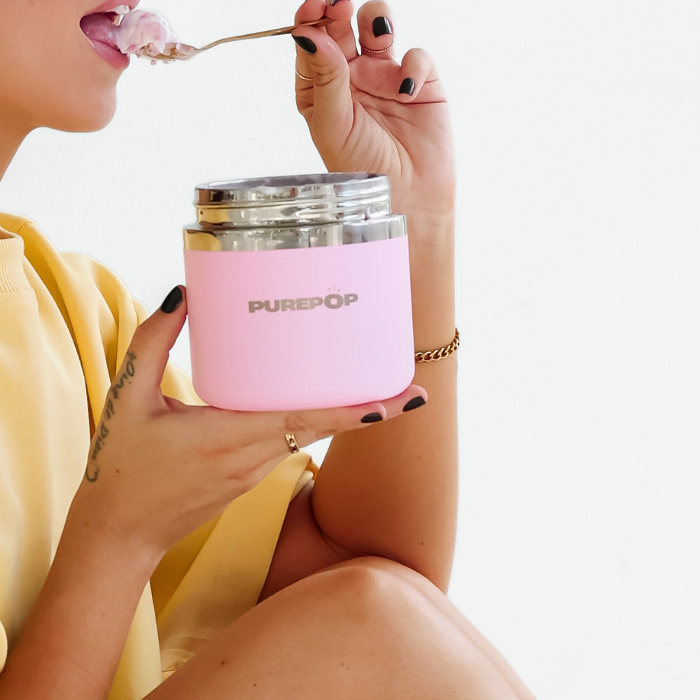 A person in a yellow shirt holds a pink PUREPOP container with a silver lid and eats with a spoon. Only the lower face and hands are visible against a white background.