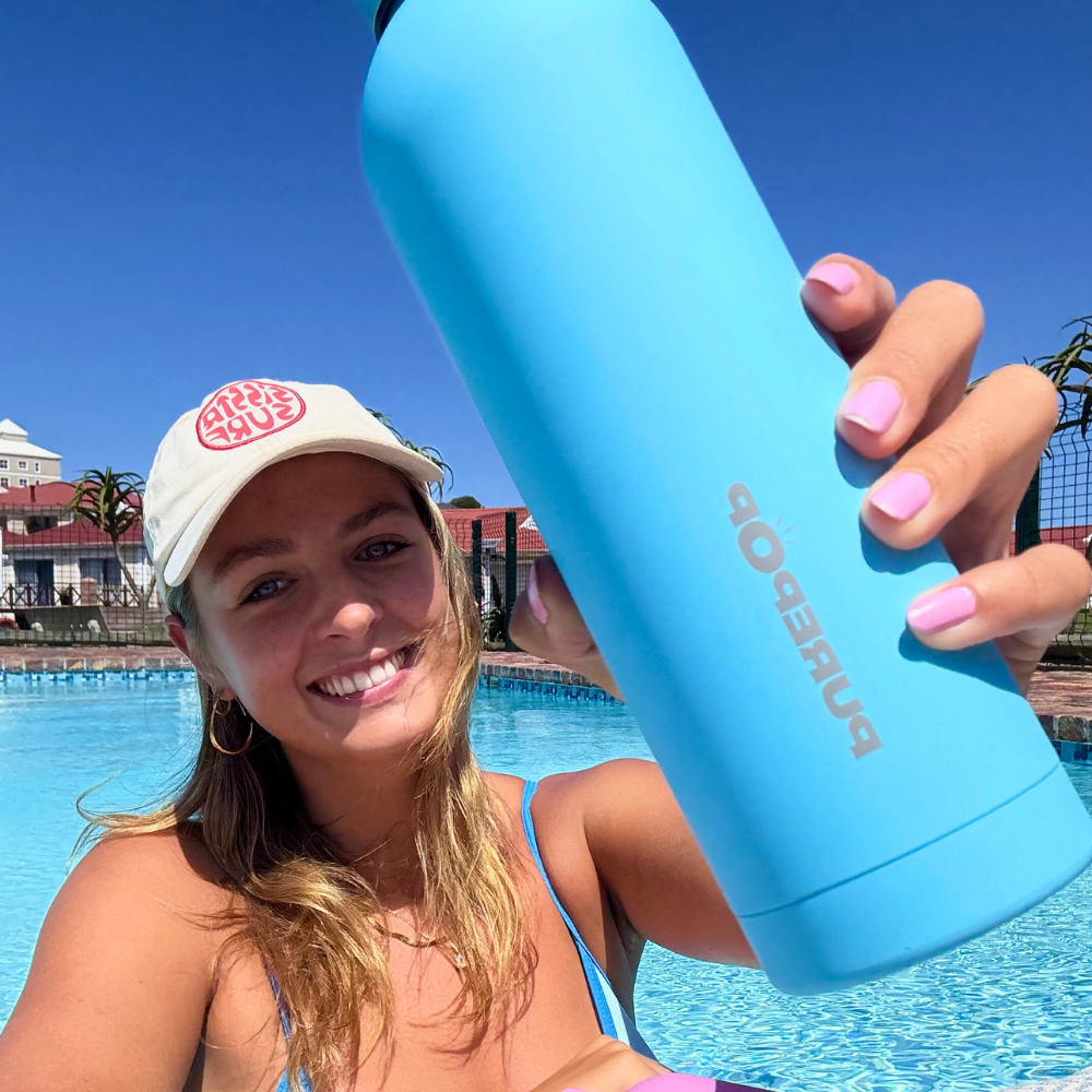 A smiling woman in a cap holds the Purepop Core Series | Frost Blue leakproof bottle towards the camera while sitting by a pool on a sunny day, with buildings and a clear blue sky in the background.