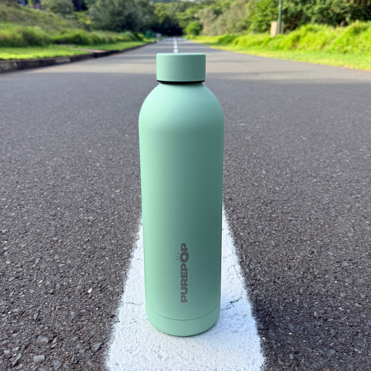 A mint green Purepop Core Series water bottle stands upright on a road’s white line, surrounded by greenery. Its leakproof design fits car cup holders, keeping your drink secure wherever you go.
