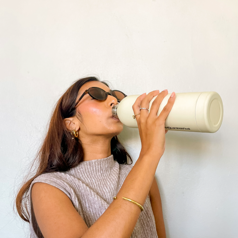 A woman wearing sunglasses and a sleeveless turtleneck top drinks from a large cream-colored bottle against a plain light background. She has long hair, statement hoop earrings, rings, and a bracelet.