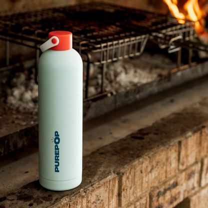 A Core Series Plus | Ice Blue double-walled stainless steel water bottle with a red cap stands on a brick surface near a grill with flames and glowing coals in the background.