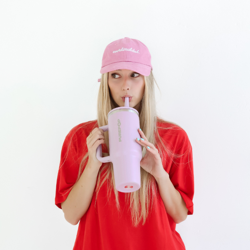 A young woman with long blonde hair, wearing a pink cap and red shirt, sips from a large lavender-colored tumbler with a straw. She stands against a plain white background.