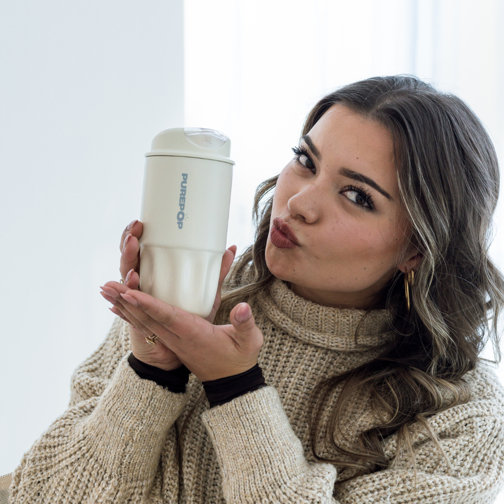 A woman in a beige sweater makes a kissing face while holding a Sip Series | Ivory PUREPOP reusable flask with a leakproof flip lid. She has long, wavy hair and large hoop earrings, set against a bright, softly lit background.