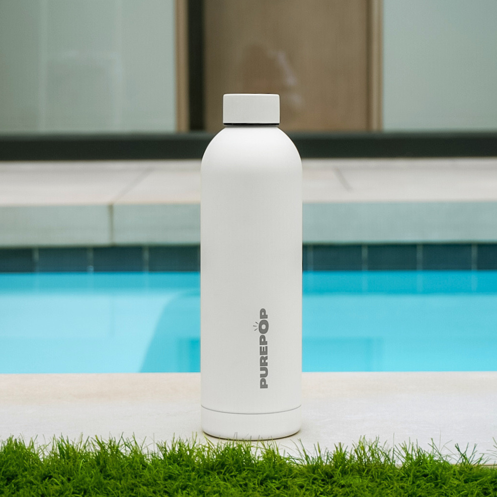 A Core Series | Frost White Purepop water bottle with thermal insulation stands upright on concrete near grass, with a swimming pool and a modern building in the background.
