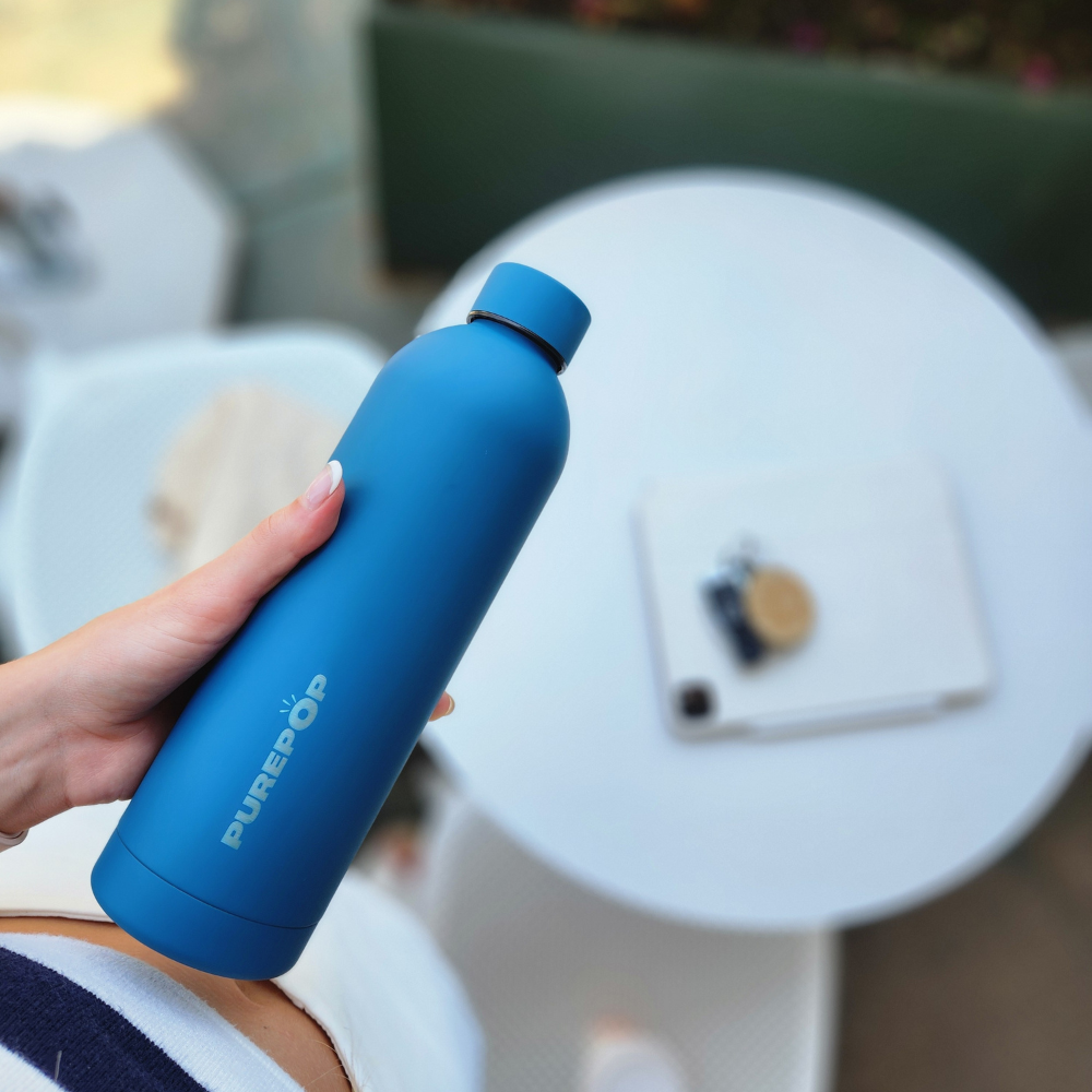 A person holds a blue PUREPÖP water bottle over a white round table with a tablet, coffee cup, and glasses on it, viewed from above, highlighting the stylish hydration companion.