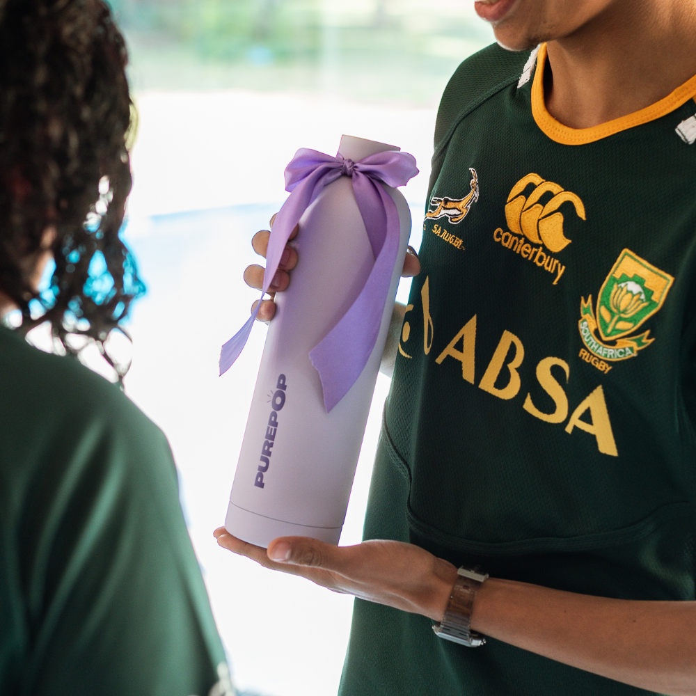 A person in a green rugby jersey hands a white Core Series Plus | Pastel Lilac flask with a purple bow to another person, both standing indoors.