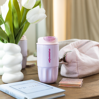 A soft lilac Sip Series leakproof travel mug sits on a table with white tulips in a vase, a textured pink bag, a blue book, and a small makeup palette in a softly lit, natural room.