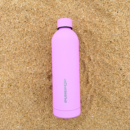 A Purepop Core Series | Pretty Pink leakproof water bottle stands upright on sandy ground, designed to fit car cup holders.