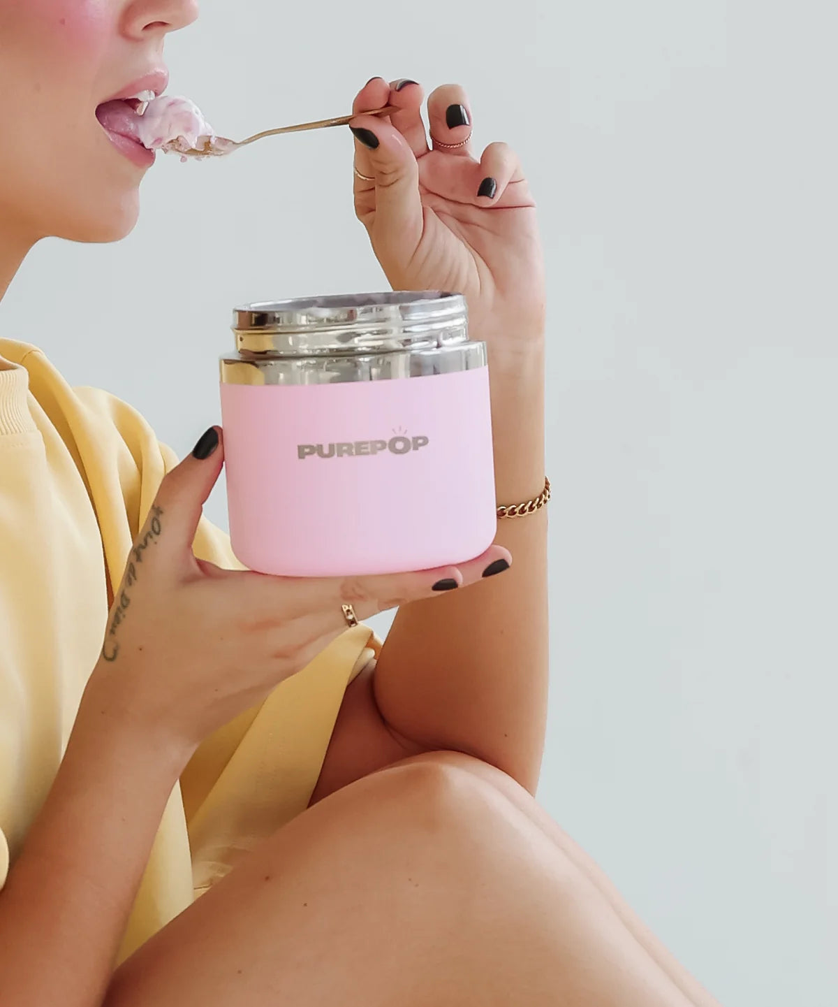 A person in a yellow top eating from an open pink PurePop Food Flask with a spoon.
