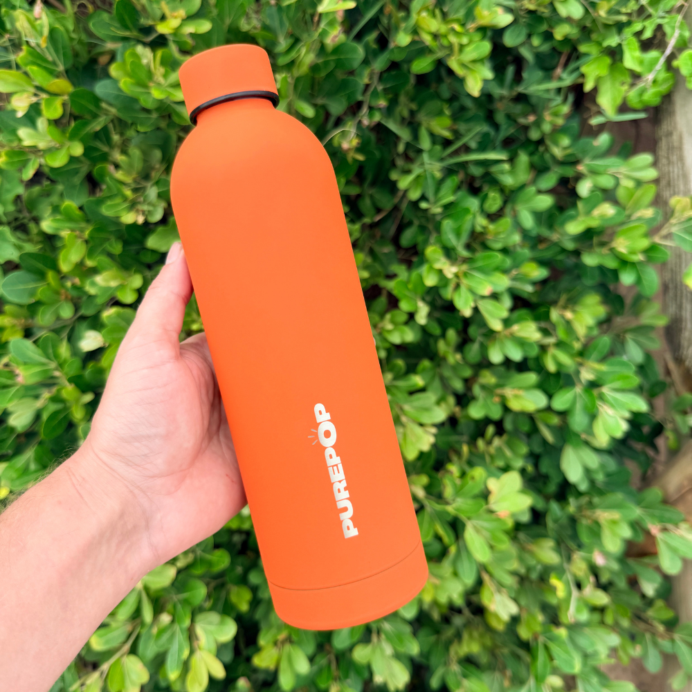 A hand holds a Sunset Orange Core Series Purepop water bottle in front of green leafy bushes.