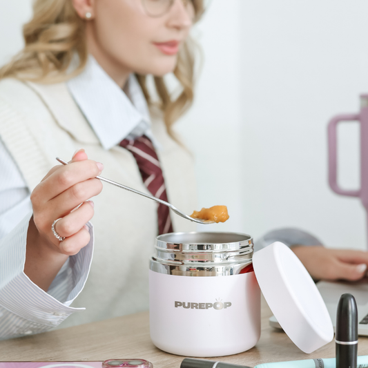 A woman in a white vest and striped tie uses a spoon to scoop food from a pink Food Flask | Frost White while sitting at a desk with various items.